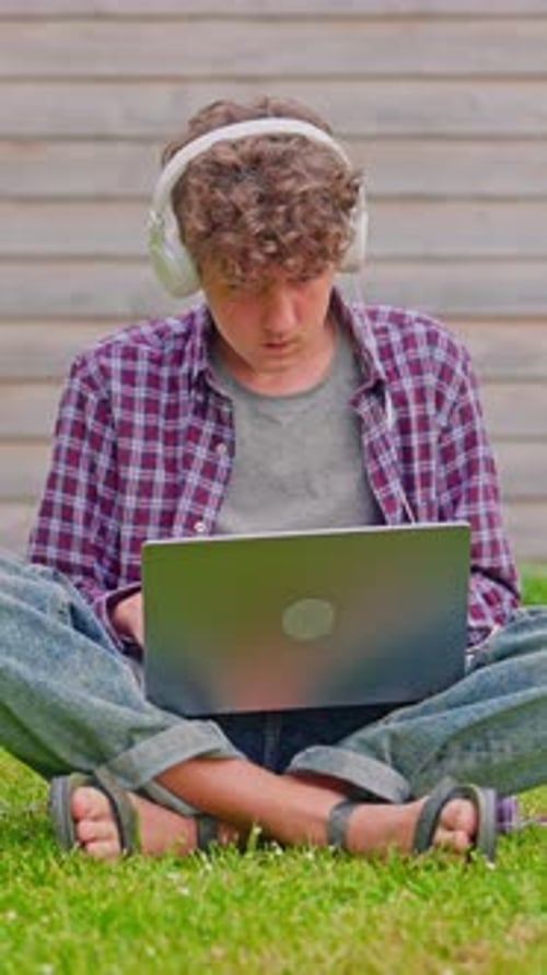 Vertical Video Curly Teenage Boy Listening to Music on Headphones and Works on a Laptop While