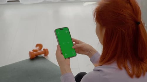 Woman looking at green screen smartphone while taking a gym break on mat