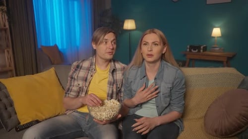 A Man and a Woman are Watching a Horror Movie While Sitting on the Couch with a Plate of Popcorn The
