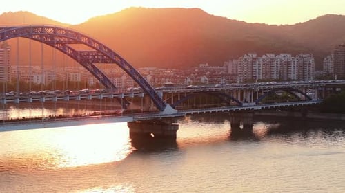 Luftaufnahme einer blauen Brücke mit Autos, die bei Sonnenuntergang einen Fluss überqueren, mit einer Skyline der Stadt in der