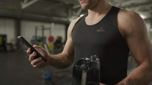 Close-Up of Muscular Man Holding Water Bottle and Using Smartphone in Gym