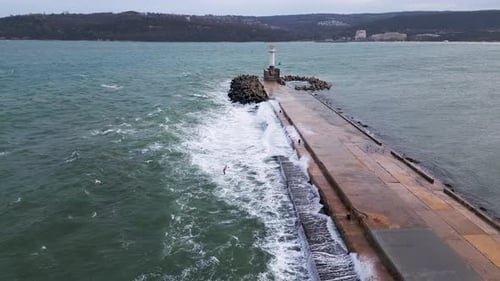Aerial View of a Long Concrete Pier with Crashing Waves and a Lighthouse at the End Surrounded By