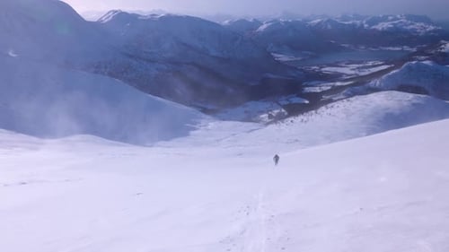Man walking uphill in the middle of a snowstorm, surrounded by beautiful mountain scenery, slight ca