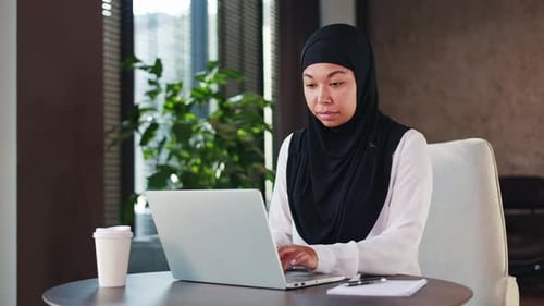 Concentrated Businesswoman in Hijab Sitting at Office Desk and Typing on Laptop