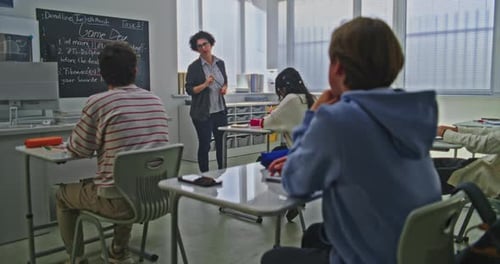 Female Teacher Stands Near Blackboard Explaining Coding Assignment to Group of Students in Bright
