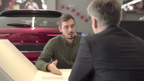 man with beard in car dealership speaks with manager with car on background
