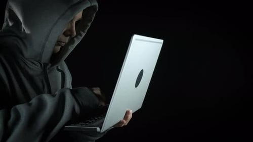 Hooded Figure Typing on Silver Laptop in Dark