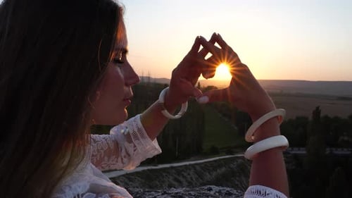 Fröhliche Frau in weißem Boho-Kleid bastelt Herzzeichen mit Händen auf Sonnenuntergang in den Bergen, romantische Frau