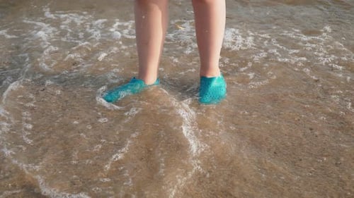 Sea Waves Wash Feet of Child in Protective Waterproof Shoes on Sand of Beach