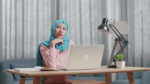 Mulher muçulmana pensando em algo enquanto trabalha em um laptop em casa