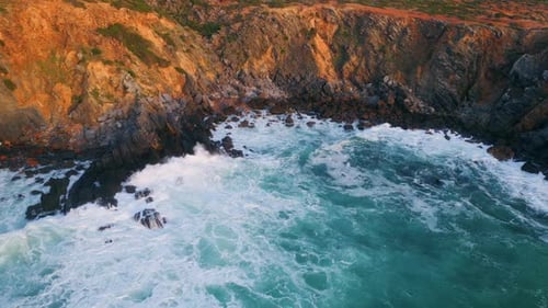 Stormy ocean waves crashing on rocky coastline in slow motion aerial view sunny day