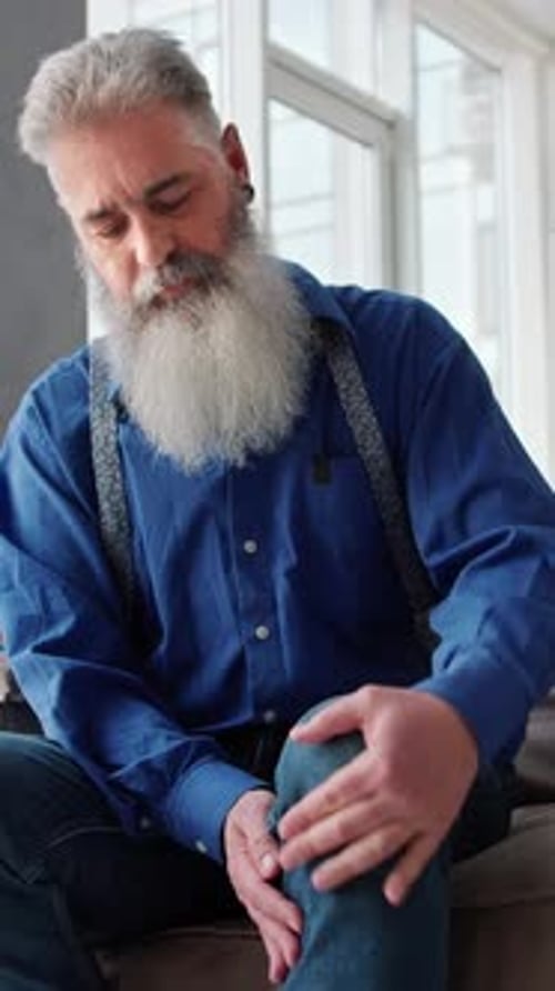 Mature Man with a Large Gray Beard Massaging His Knee on the Sofa at Home