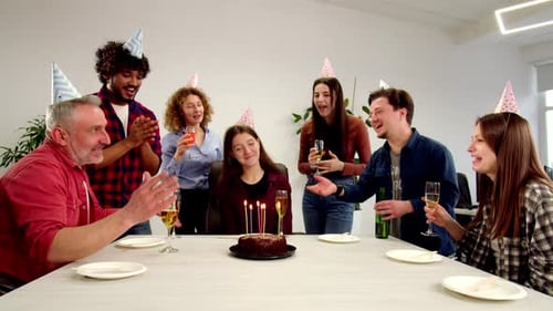A Brunette Woman Sits at a Table in a Circle of Friends and Blows Out Candles on a Chocolate Cake