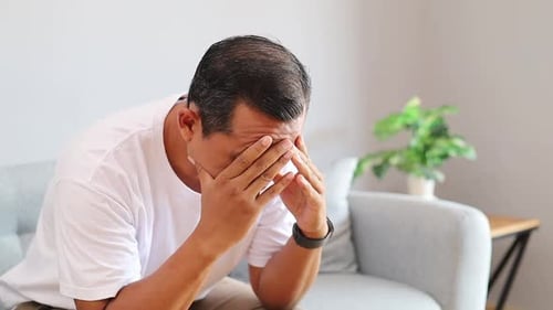 Stressed Asian man sitting on sofa with hands covering face showing emotional struggle anxiety and