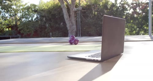 Close up of yoga mat, laptop, bottle with juice and weights on floor at sunny home, slow motion