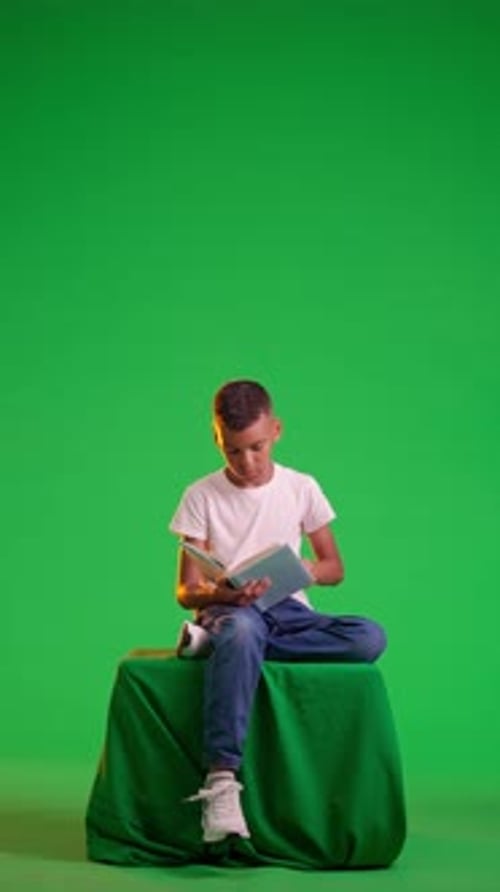 Young Boy Reading a Book Attentively While Seated Against on a Green Screen Chroma Key