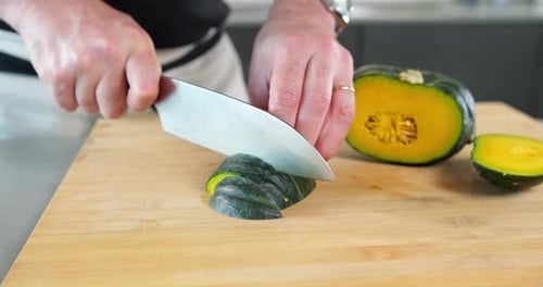 Male chef roughly chopping big slices of green pumpkin on the kitchen with the sharp knife