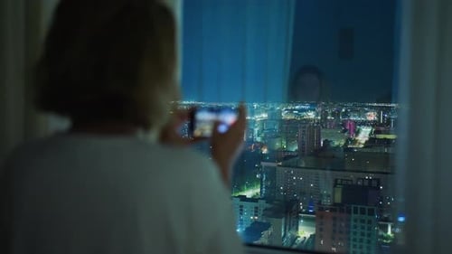 Woman Taking Pictures of Illuminated Cityscape at Night From Hotel Room