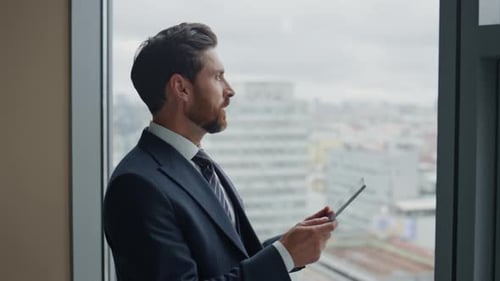 Successful bearded businessman rejoicing company achievement holding tablet near office window