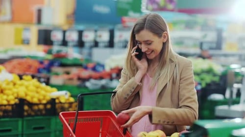 Attractive Young Woman Talking on Phone Smiling Chooses Apples and Puts in Basket in Supermarket