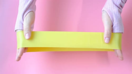 Elástico de fitness que alonga as mãos em close-up de fundo rosa.