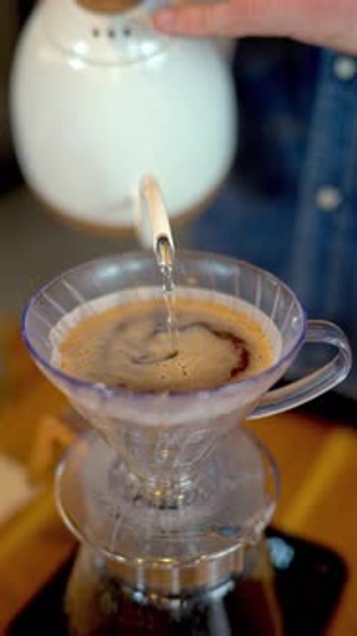 Close up of a man brewing pour-over drip coffee in a slow, circular motion on a wooden table. Vertic