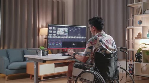 Back View Of Asian Man In Wheelchair Is Using A Desktop Editing The Video And Color Grading At Home