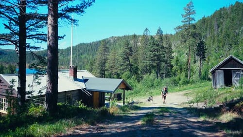 Portrait Of Backpacker With His Dog Near Mountain Cabin During Summer. Wide Shot