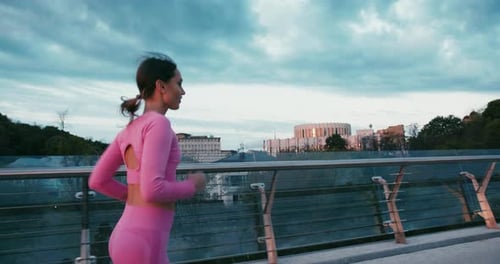 Young Woman in Pink Sportswear Runs on Bridge Towards Sunset