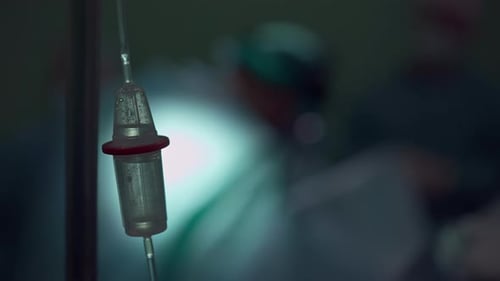Nahaufnahme des Tropfenzählers im Dunkeln mit verschwommenem Chirurgen, der im Hintergrund eine Operation durchführt