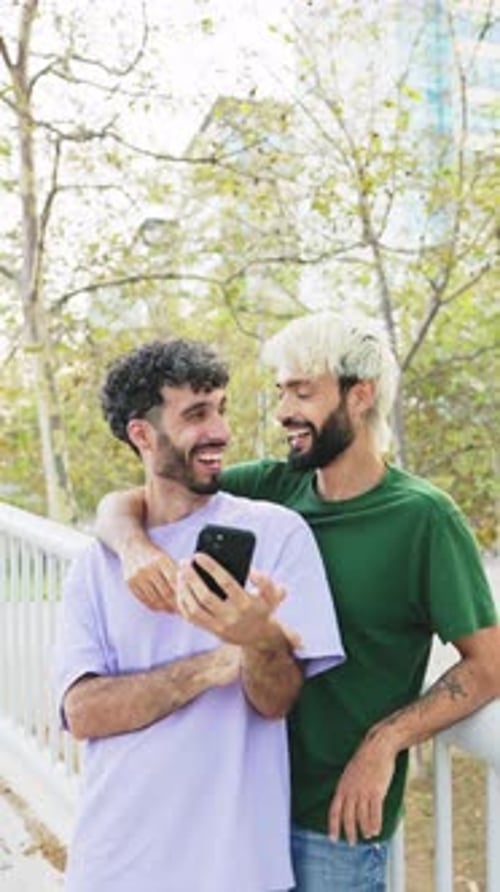 Vertical video of a cheerful gay couple using mobile phone in a park in daylight.