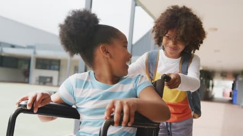 Video of two happy diverse schoolchildren, boy pushing girl in wheelchair in corridor