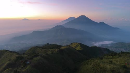 Aerial sunrise view of misty mountain ranges and volcanic peaks from lush green hills.