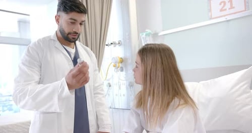 Diverse male doctor showing girl patient in hospital bed how to use asthma inhaler, slow motion