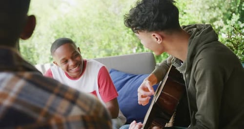 Happy diverse male teenage friends playing guitar at home, slow motion