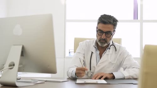 Male Doctor Noting Something on Document and Using Computer in Clinic
