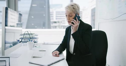 4k video footage of a mature businesswoman writing notes while talking on a cellphone in an office
