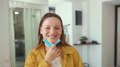 Portrait of Young Woman Removes Medical Mask Face in Coronavirus Pandemic at Home Looking at Camera