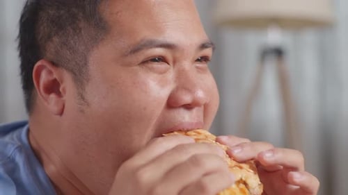 Close Up Of A Fat Asian Man Eating Fast Food Having A Hamburger On A Sofa In The Living Room At Home