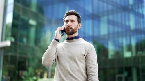 Man smiling while talking on the phone in an urban setting near a modern office building.