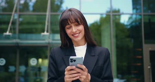 Close up of a smiling businesswoman in formal attire walking outdoors and using smartphone.