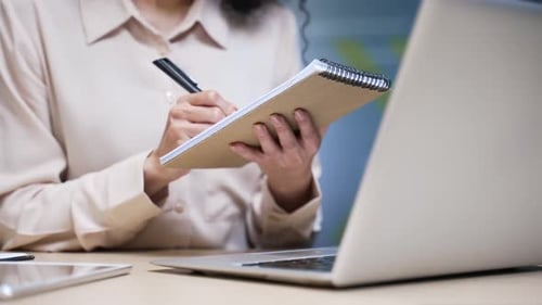 Nahaufnahme der Hände einer Geschäftsfrau, die Notizen im Notizbuch macht, während sie den Laptop am Schreibtisch benutzt