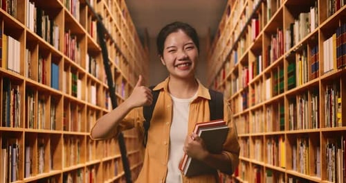 Asian Woman Student With A Backpack And Some Books Smiling And Showing Thumbs Up In The Library