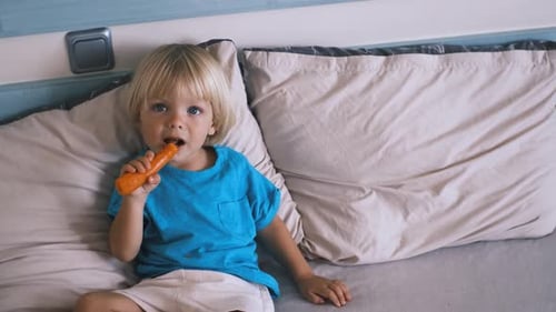 Blonde Haired Child Eating Carrot on a Bed