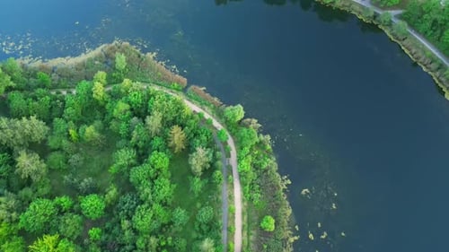 Drone flight over the banks of a narrow lake, with a view of the water, trees, and bike paths