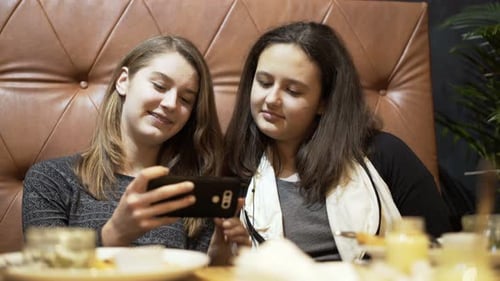 Teenage girlfriends enjoying funny movie on smartphone screen at cafe table