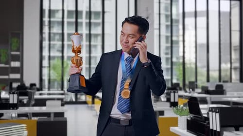 Asian Business Man With A Gold Medal Holding A Gold Trophy And Talking On Smartphone In Office