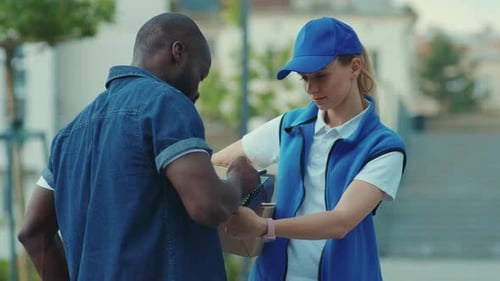 Young Woman Courier in Blue Hat Hold Box for African American Client Paying By Phone for Parcel