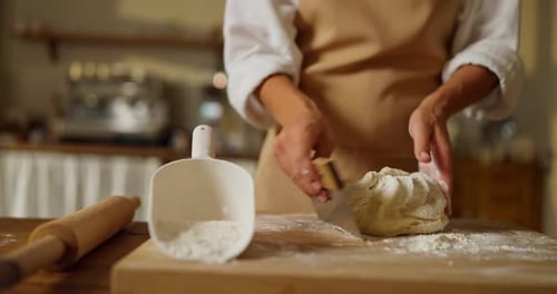 Girl Baker in a White Shirt and Beige Apron Kneads Dough Using a Pastry Spatula on a Cutting Board
