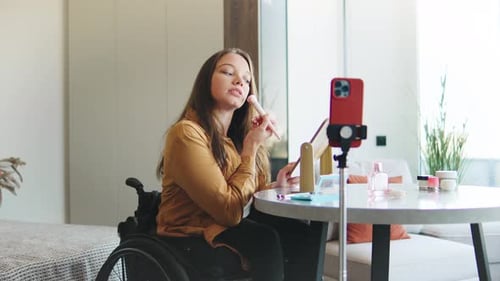 Mujer sonriente en silla de ruedas maquillándose mientras usa el teléfono inteligente para un tutorial en vídeo en un